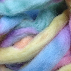 May include: Close-up of colorful fiber strands in pastel hues. The image features strands of yellow, blue, green, purple, and peach. The fibers appear soft and fluffy, suggesting a textile or craft material. The colors blend together, creating a visually appealing texture.