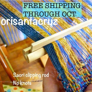 May include: A wooden Saori clipping rod used for weaving, featuring a slotted design to hold warp threads without knots. The rod is positioned against a loom with blue and gold yarn threads, with the text Saorisantacruz and Saori clipping rod No knots.