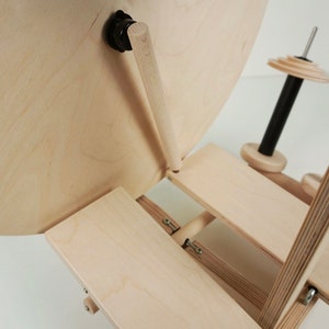 Puede incluir: Un torno de hilar de madera con una base grande, redonda, de madera clara y una rueda m&aacute;s peque&ntilde;a, redonda, de madera clara. El torno de hilar est&aacute; plegado y la base descansa sobre una plataforma de madera clara. El torno de hilar tiene un husillo de metal negro y un mango de madera clara.