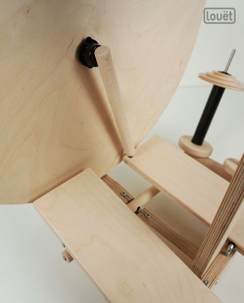 Puede incluir: Un torno de hilar de madera con una base grande, redonda, de madera clara y una rueda m&aacute;s peque&ntilde;a, redonda, de madera clara. El torno de hilar est&aacute; plegado y la base descansa sobre una plataforma de madera clara. El torno de hilar tiene un husillo de metal negro y un mango de madera clara.