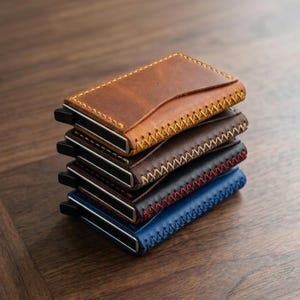 Op de afbeelding: Een stapel van vijf leren portemonnees in verschillende kleuren: bruin, donkerbruin, zwart en blauw. Elke portemonnee heeft een gestikte rand en een metalen geldclip. De portemonnees liggen op een houten oppervlak.