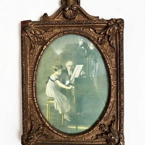 May include: An ornate, rectangular, antique-style gold frame with an oval center opening. The frame features intricate relief carvings at the corners and top. Inside is a print of a young person in a white dress playing piano with an older person.