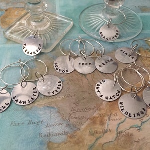 Puede incluir: Encantos de vino plateados con nombres estampados como "Targaryen", "Baratheon", "Stark", "Lannister", "Martell", "Frey", "Arryn", "Tyrell", "Night's Watch", "Wildlings" y "Greyjoy".