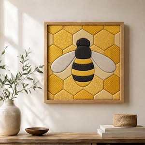 Op de afbeelding: Een vierkant wandkunstwerk met een met stof bekleed honingraatpatroon in tinten geel en goud. Een centraal bijenmotief is opgebouwd uit zwarte, gele en lichtgrijze stofsegmenten, omlijst door een lichtgekleurde houten rand.