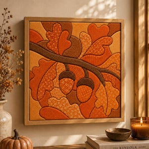 May include: A square wall art piece featuring a fabric-stitched design of oak leaves and two acorns on a branch. The artwork uses a warm palette of orange, brown, and gold tones with various floral and dotted patterns, all encased in a light wood frame.