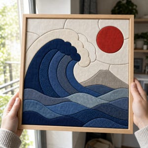 May include: A square fabric wall art piece in a light wood frame featuring a quilted design of a large blue ocean wave, a beige mountain, and a solid red sun against a cream background, constructed from various shades of blue patterned fabric.