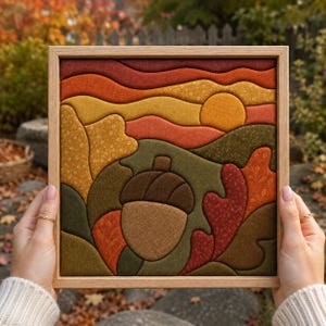 May include: A square framed fabric wall art piece featuring a layered landscape design. The scene depicts a central brown acorn surrounded by stylized autumn leaves in shades of red, orange, and olive green, with a golden sun against wavy hills.