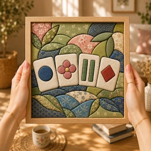 May include: A square framed textile art piece featuring four off-white rectangular tiles with raised fabric symbols: a blue circle, a pink flower, two green vertical bars, and a red diamond. The background consists of quilted fabric patches in shades of green, blue, and pink with floral and geometric patterns.