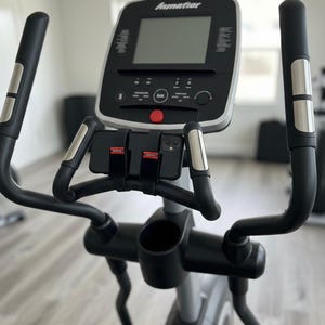 May include: A black and silver elliptical machine with a digital display and multiple control buttons. The machine has black handles with silver accents and a built-in phone holder. The brand name "Ammator" is visible on the display.