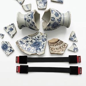 Puede incluir: Fragmentos dispersos de un jarrón y platos antiguos de porcelana rota, con diseños florales azules sobre fondo blanco. Dos correas negras con detalles rojos y el texto "ORIGINAL" son visibles. Las piezas están dispuestas sobre una superficie blanca.