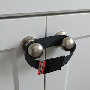 May include: A black cabinet door lock with a red and white label that reads "Snug & Spils". The lock is secured around two silver door knobs on a white cabinet.