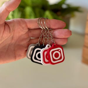 Op de afbeelding: Een set van vijf sleutelhangers, elk met een vierkant ontwerp met een camera-icoon. De sleutelhangers zijn grijs, zwart, wit en rood, met een zilveren sleutelring. De sleutelhangers worden in een hand gehouden.