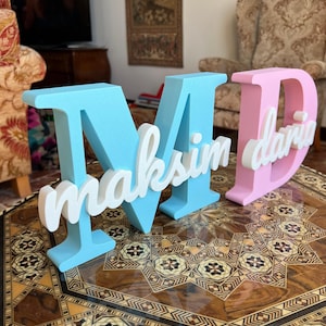 Può includere: Lettere decorative in legno che compongono "MD" in azzurro e rosa, con i nomi "maksim" e "daria" in corsivo bianco. Le lettere sono esposte su un tavolo decorativo in legno.