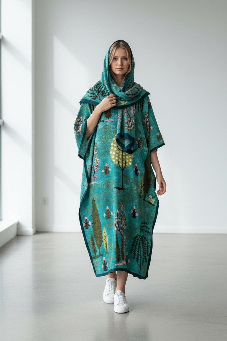 Puede incluir: Un caft&aacute;n con capucha de color turquesa con un estampado de tem&aacute;tica natural con &aacute;rboles y flores. El caft&aacute;n tiene un borde turquesa oscuro y se combina con una bufanda a juego. La persona lleva zapatillas blancas.