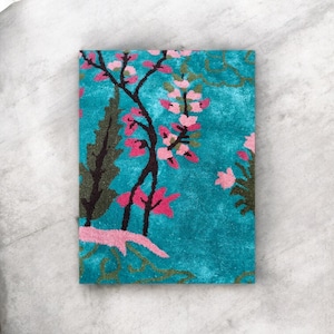 Puede incluir: Una alfombra rectangular turquesa con un dise&ntilde;o floral. La alfombra presenta un &aacute;rbol con flores rosas y hojas verdes oscuras. El fondo es de color turquesa s&oacute;lido, y el dise&ntilde;o general recuerda a un jard&iacute;n.