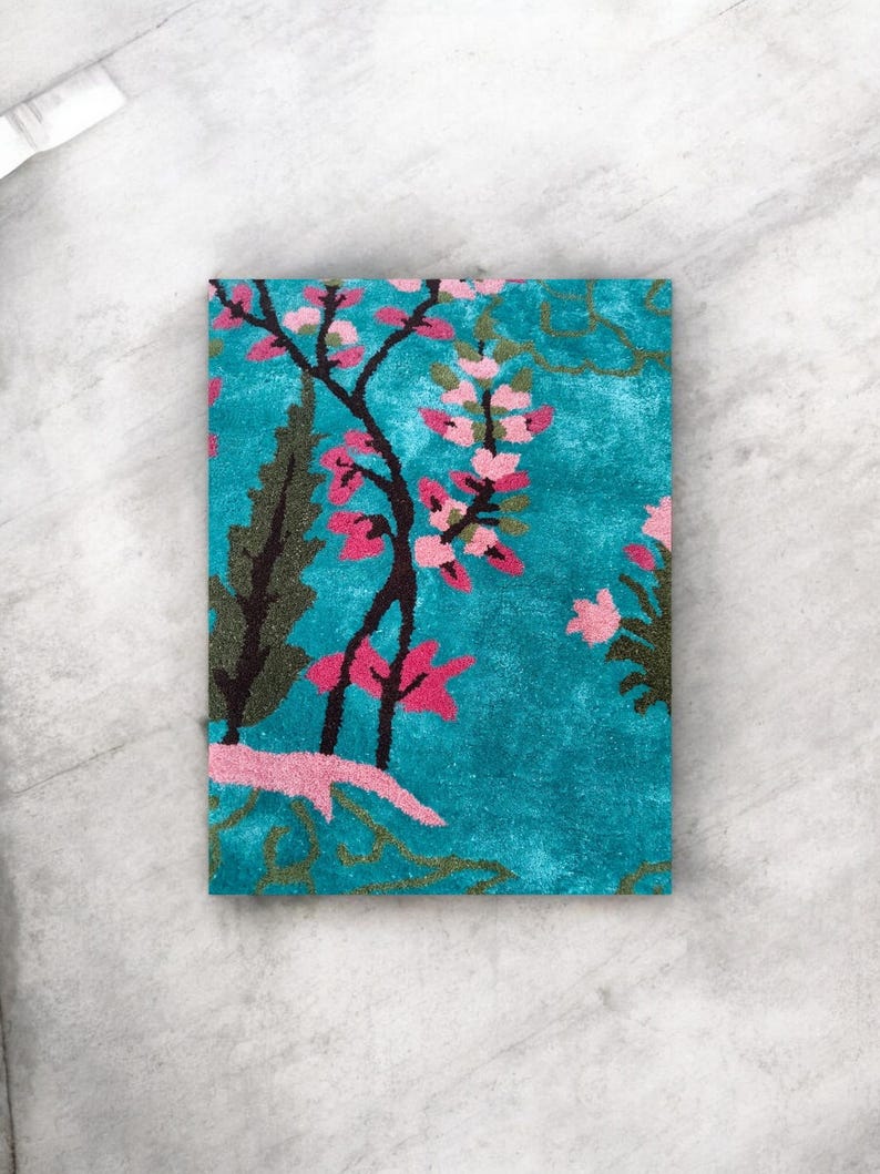 Puede incluir: Una alfombra rectangular turquesa con un dise&ntilde;o floral. La alfombra presenta un &aacute;rbol con flores rosas y hojas verdes oscuras. El fondo es de color turquesa s&oacute;lido, y el dise&ntilde;o general recuerda a un jard&iacute;n.