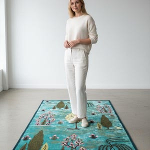 Puede incluir: Una alfombra rectangular turquesa con borde azul oscuro. La alfombra presenta un dise&ntilde;o de paisaje con &aacute;rboles, flores y un estanque. La alfombra mide aproximadamente 152 cm de largo y 91 cm de ancho.