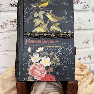 May include: A black book cover featuring a vintage botanical illustration of two birds perched on branches, white and pink roses, and cursive script text. The text reads J-STECKLER SEED CO. LTD. SOUTHERN STATES. A decorative gold-toned metal trim runs horizontally.