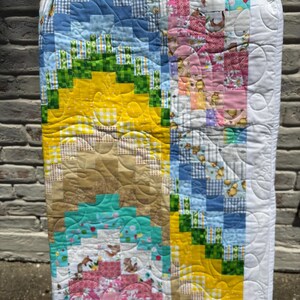 May include: A handmade quilt with a patchwork design. The quilt features a variety of fabrics in shades of blue, yellow, green, pink, and brown. The quilt has a white border and is hanging against a brick wall.