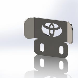 Puede incluir: Un soporte metálico gris con el logotipo de Toyota recortado. El soporte tiene forma rectangular con esquinas redondeadas y dos orificios ovalados en la parte inferior. El logotipo de Toyota está en el centro.