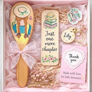 Puede incluir: Una caja de regalo con un cepillo de pelo de madera, un marcapáginas, un llavero, pegatinas y una tarjeta. El cepillo de pelo presenta a una niña con trenzas y un sombrero de paja. El marcapáginas dice "Just one more chapter". El llavero lleva grabado el nombre "Lily".