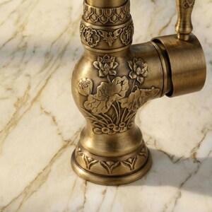 May include: Close-up of an antique brass faucet with intricate floral and oriental design. The faucet features detailed carvings of lotus flowers and leaves, with a warm, aged finish. The handle and base also have decorative patterns.
