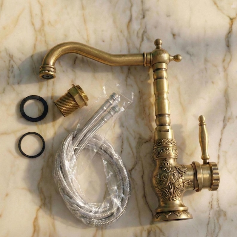 May include: An antique-style brass kitchen faucet with a curved spout and ornate floral detailing. Includes flexible supply lines, a threaded connector, and two black rubber washers. The faucet has a lever handle and a decorative top.