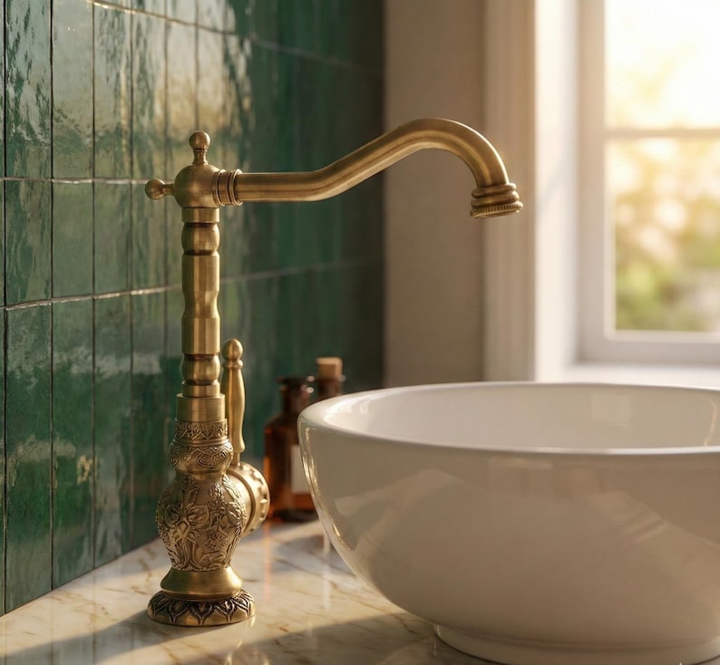 May include: A gold-colored faucet with an ornate design, featuring a curved spout and detailed base. The faucet is set against a backdrop of green tiles and a white sink, suggesting a bathroom setting.