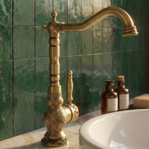 May include: Antique brass faucet with an ornate design. The faucet features a curved spout and a decorative handle. The base has an engraved floral pattern. The faucet is set against a green tiled wall and a white sink.