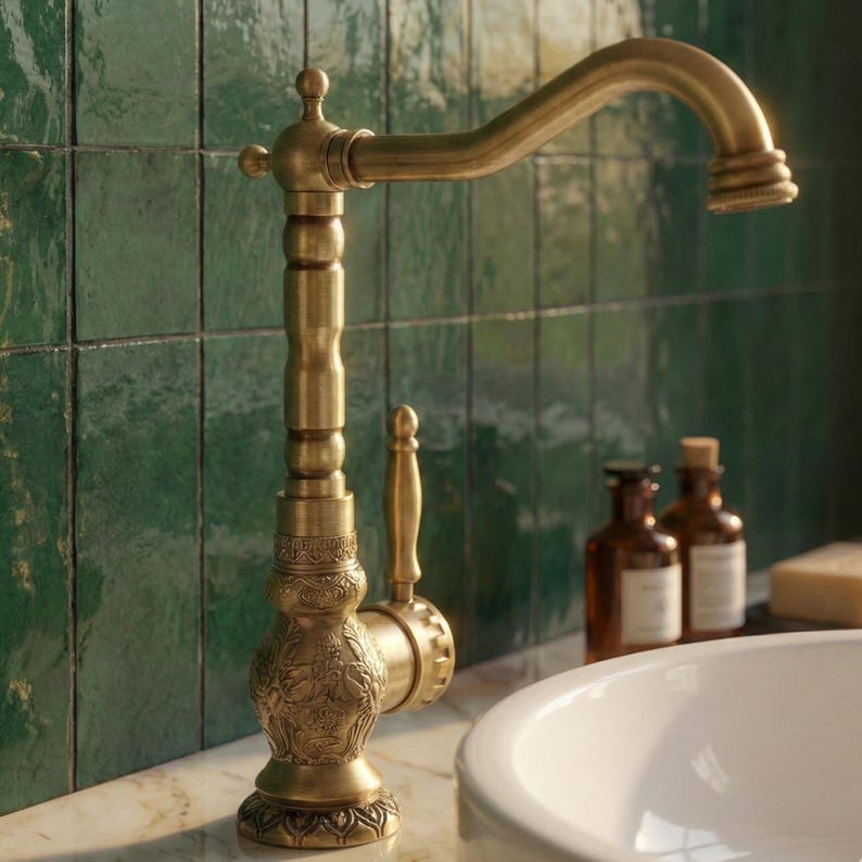 May include: Antique brass faucet with an ornate design. The faucet features a curved spout and a decorative handle. The base has an engraved floral pattern. The faucet is set against a green tiled wall and a white sink.
