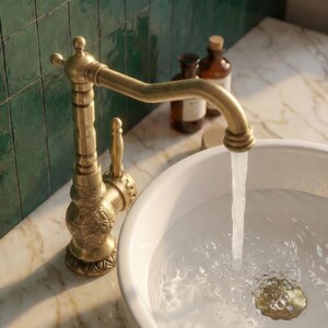 May include: An antique-style brass faucet with intricate floral detailing is shown, with water flowing into a white ceramic sink. The faucet has a curved spout and a lever handle. The sink is set on a marble countertop.