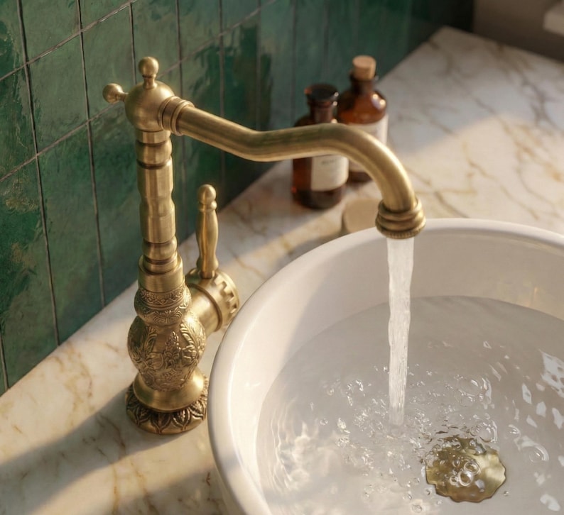 May include: An antique-style brass faucet with intricate floral detailing is shown, with water flowing into a white ceramic sink. The faucet has a curved spout and a lever handle. The sink is set on a marble countertop.