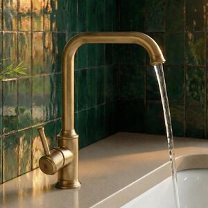 May include: A brass-coloured kitchen tap with a curved spout, pouring water into a sink. The tap is set against a backdrop of green tiles. A glass bottle with a plant is on the left.