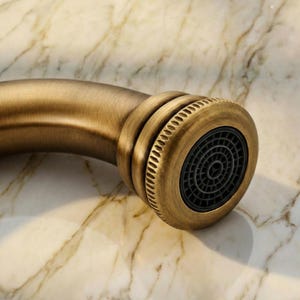 May include: Close-up of a bronze-colored faucet head with a black aerator. The faucet has a curved spout and a decorative, ridged design around the head. The background is a marble-like surface.