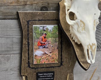 May include: A wooden plaque shaped like the state of Montana featuring a mounted deer skull, a photograph of a hunter with a deer, and a black nameplate reading Swan Mountains Carvers & Crafters and SWANMTNSCNC@GMAIL.COM with barbed wire art.