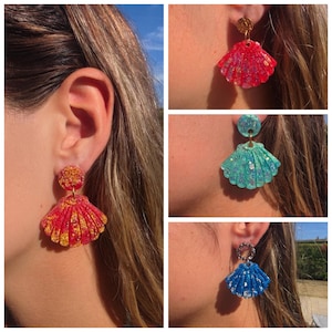 Peut inclure: Une collection de quatre boucles d'oreilles pendantes en forme de coquillages, chacune composée d'un clou d'oreille circulaire et d'un pendentif en forme d'éventail. Elles sont disponibles en rouge avec des éclats dorés, turquoise pailleté et bleu avec des reflets argentés.