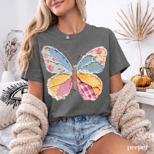 May include: A charcoal grey t-shirt with a patchwork butterfly design. The butterfly is made of various fabrics in blue, yellow, pink, and red. The t-shirt is a relaxed, casual style.