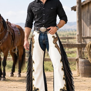 May include: A pair of white and black leather chaps featuring long fringe along the sides and intricate gold-toned floral embroidery. The chaps are worn over dark blue denim jeans, paired with a black long-sleeved button-down shirt and a brown hat.