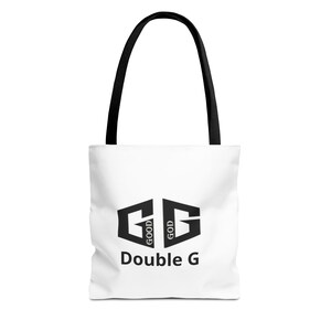 Puede incluir: Bolso tote blanco con asas negras. Presenta un logotipo Double G negro con las palabras "GOOD GOD" a cada lado de las G. Las palabras "Double G" están impresas debajo del logotipo.