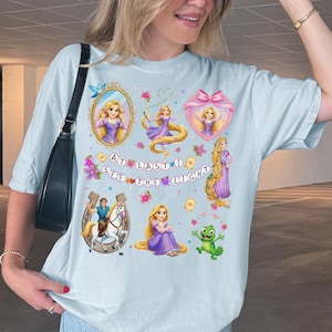May include: A light blue t-shirt featuring a graphic print of Rapunzel, a horse, and a chameleon. The design includes various illustrations of the character, floral accents, and the text AT LAST I SEE THE LIGHT in circular bubbles across the center.