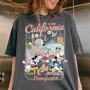 Puede incluir: Camiseta gris oscuro con un gr&aacute;fico de Disneyland. El gr&aacute;fico incluye el texto "California Est. 1955" e im&aacute;genes de Mickey Mouse, Minnie Mouse, Donald Duck y otros personajes de Disney frente a las atracciones del parque.