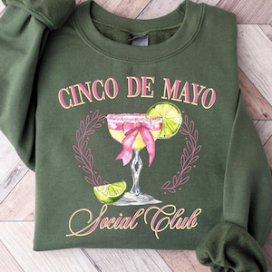 Puede incluir: Una sudadera verde oliva con las palabras "Cinco de Mayo Social Club" en rosa. El diseño presenta una margarita con un lazo rosa y una rodaja de lima. La sudadera está doblada sobre una superficie de madera clara.