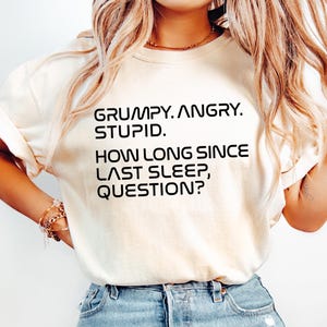 May include: Cream-colored t-shirt with the text "GRUMPY. ANGRY. STUPID. HOW LONG SINCE LAST SLEEP, QUESTION?" in black. The shirt is paired with light blue denim jeans.