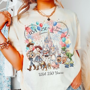 Peut inclure: Un t-shirt couleur cr&egrave;me avec un motif repr&eacute;sentant divers personnages anim&eacute;s, un ch&acirc;teau, des feux d'artifice et des ballons. Le texte indique USA 250 Years Celebration et USA 250 Years. Le design inclut Woody, Jessie, Buzz l'&Eacute;clair et autres.