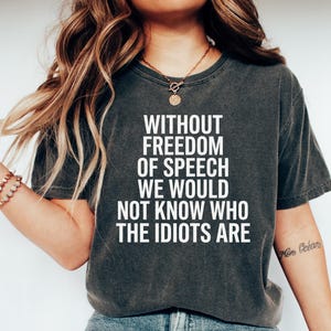 May include: A charcoal grey short-sleeved t-shirt featuring white text that reads: WITHOUT FREEDOM OF SPEECH WE WOULD NOT KNOW WHO THE IDIOTS ARE. The shirt has a crew neckline and a vintage-style washed finish, paired with blue denim jeans.