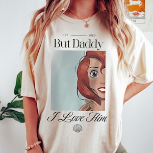 Puede incluir: Camiseta color crema con el texto "But Daddy I Love Him" y un dibujo animado. La camiseta tiene una est&eacute;tica vintage, con el a&ntilde;o "1989" impreso encima del texto. El dise&ntilde;o incluye un gr&aacute;fico de concha.