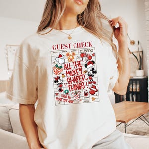 Pode incluir: Uma t-shirt de cor creme com um gráfico vermelho e preto de uma conta de restaurante. O texto diz GUEST CHECK, ALL THE MICKEY SHAPED THINGS e THANK YOU PLEASE COME AGAIN, rodeado por vários ícones do Mickey Mouse com tema natalício.