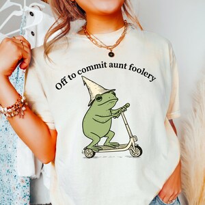 May include: A cream-colored t-shirt featuring a graphic of a green frog wearing a pointed hat and riding a kick scooter. The text above the frog reads, Off to commit aunt foolery, printed in a black, curved, sans-serif font on the front center.