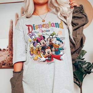 K&ouml;nnte beinhalten: Ein graues T-Shirt mit einem farbenfrohen Aufdruck von Disney-Figuren, darunter Mickey Mouse, Minnie Mouse, Donald Duck und Goofy. Das Shirt tr&auml;gt den Text "Disneyland 2026" in einer Regenbogen-Schriftart. Das Design beinhaltet Unterschriften und Feuerwerk.