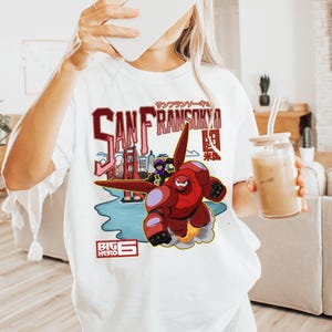 May include: A white short-sleeved t-shirt featuring a graphic print of the character Baymax from Big Hero 6 flying over a stylised San Fransokyo cityscape. The design includes red and black text, the Golden Gate Bridge, and the Big Hero 6 logo.